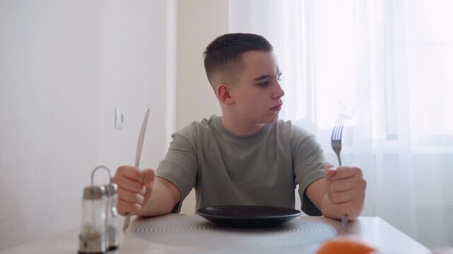Hungry child sits at dining table holding spoon and fork, tapping utensils on empty plate with impatient expression while looking to side, waiting for meal in bright home kitchen filled with daylight