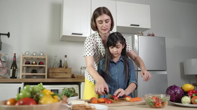 Smart caucasian mother and asian girl cooking together and chopping vegetable or preparing salad for dinner. Happy mom and daughter making healthy food with fresh food. Healthy food concept. Pedagogy.