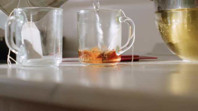 Close up of hot water being poured into glass cup with teabag inside, creating rich amber color as tea steeps beside mixing bowl in bright kitchen setting during homemade drink preparation