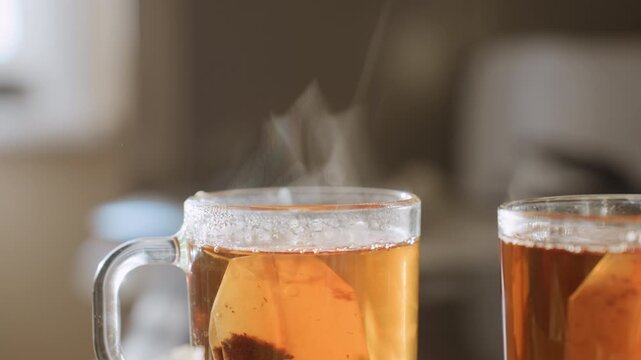 Close up of steaming herbal tea in transparent glass cup with teabag inside, vapor rising gracefully from hot surface, condensation forming around rim, warm amber liquid glowing in soft kitchen light