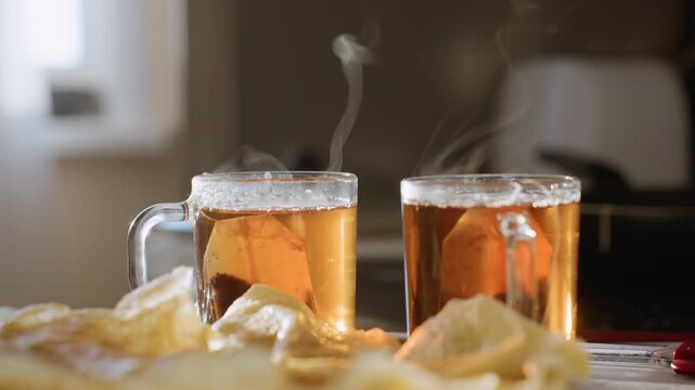 Close up of two steaming glass cups filled with hot herbal tea and teabags inside, vapor rising above surface, pancakes served on plate in foreground, cozy breakfast setting
