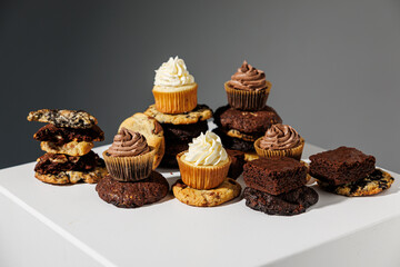 Artistic arrangement of chocolate and vanilla cupcakes, stacked cookies, and fudgy brownies on a white pedestal with a gray backdrop and copy space