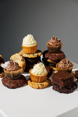 Artistic arrangement of chocolate and vanilla cupcakes, stacked cookies, and fudgy brownies on a white pedestal with a gray backdrop and copy space