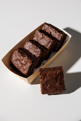 A row of rich, chocolate brownies in a kraft paper tray with one piece placed in front on a white surface, lit by strong natural light with copy space