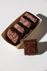 A row of rich, chocolate brownies in a kraft paper tray with one piece placed in front on a white surface, lit by strong natural light with copy space