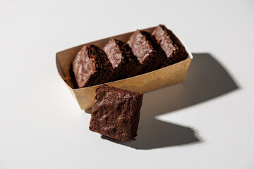 A row of rich, chocolate brownies in a kraft paper tray with one piece placed in front on a white surface, lit by strong natural light with copy space