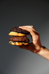 A hand grips a tall stack of four assorted cookies in different flavors, against a soft gradient gray background with dramatic lighting and copy space
