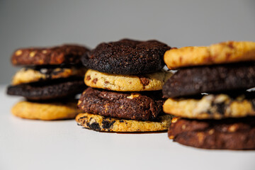 Close-up image of stacked cookies in various flavors including chocolate chip, double chocolate, and cookies and cream, arranged on a white surface with a soft gray background