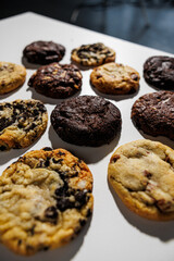 Top-down view of assorted cookies including chocolate chip, double chocolate, white chocolate chunk, and cookies and cream, laid out in a grid on a white background with copy space