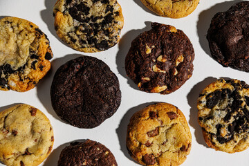 Top-down view of assorted cookies including chocolate chip, double chocolate, white chocolate chunk, and cookies and cream, laid out in a grid on a white background with copy space