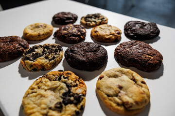 Top-down view of assorted cookies including chocolate chip, double chocolate, white chocolate chunk, and cookies and cream, laid out in a grid on a white background with copy space
