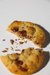 A chocolate chip cookie broken in half lies on a white surface, with scattered crumbs adding a touch of realism. No visible branding or packaging