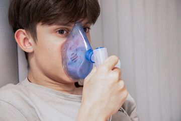 Young boy sitting in bed and using a medical nebulizer inhaler for breathing therapy. Concept of home healthcare, respiratory disease treatment, asthma, or cold recovery.