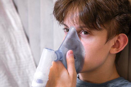 Young boy sitting in bed and using a medical nebulizer inhaler for breathing therapy. Concept of home healthcare, respiratory disease treatment, asthma, or cold recovery.
