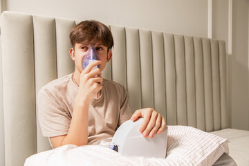 Young boy sitting in bed and using a medical nebulizer inhaler for breathing therapy. Concept of home healthcare, respiratory disease treatment, asthma, or cold recovery.