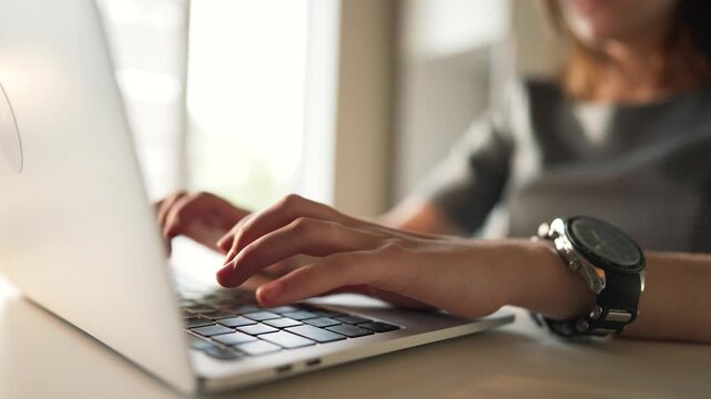 typing on laptop with hands and watch visible on wrist as woman types on keyboard at desk in office for work and business closeup of keyboard and laptop showing focused hand movement and typing