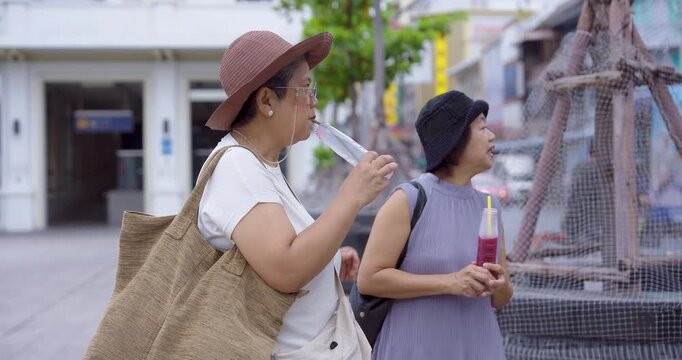 Senior asian women travelers walking on city street holding fruit drinks enjoying retirement lifestyle friendship vacation exploring culture together during urban journey relaxation happiness travel