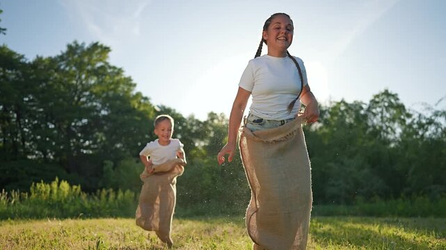 Girl wins summer sack race game. Boy jumps behind outdoors. Child enjoys fun competition in nature. Girl jumps high. Kids in summer race. Children feel joy playing outdoors competition jumping.