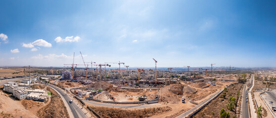 Aerial view of a sprawling construction site where red cranes stand tall against the backdrop of a city under a vast blue sky, El'ad, Center District, Israel.