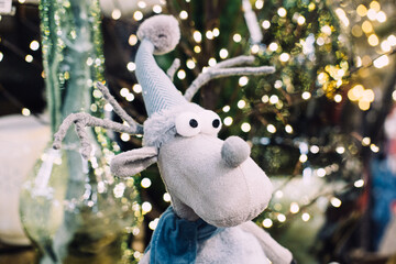 A soft plush reindeer toy with a long striped hat and googly eyes set against a background of Christmas lights. Quirky Christmas decor, playful reindeer toy, whimsical holiday trends