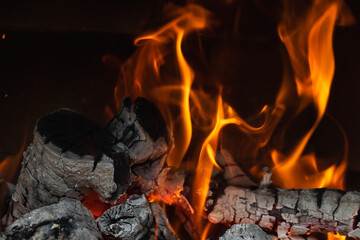 Close-up of blazing orange flames and glowing red embers on burning firewood logs in a dark, cozy hearth.