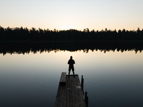 Aerial drone view of a person's silhouette on a wooden pier overlooking a tranquil Estonian lake with perfect forest reflections at sunset.