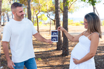 Pregnant couple holding ultrasound image of future baby outdoors. Expecting parents smiling with joy and tenderness, representing love, anticipation, and family connection during pregnancy in nature.