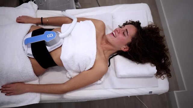 Top down view of a smiling young woman undergoing a non invasive body contouring treatment, with a high intensity electromagnetic device stimulating her abdominal muscles for fat reduction