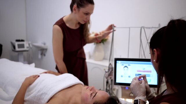 Young woman lying on a bed in a cosmetology clinic while two beauticians prepare a modern microcurrent lifting machine to perform a rejuvenating facial treatment on her skin