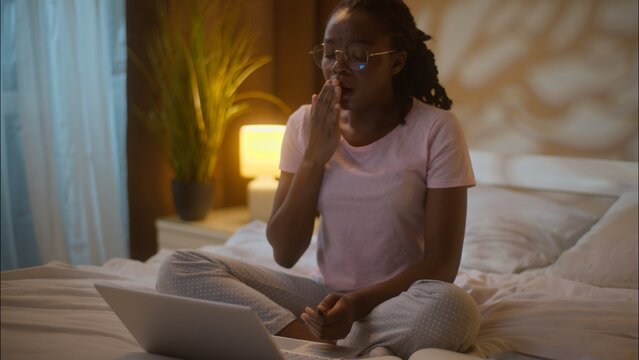 Sleepy tired African American woman student female girl working laptop computer typing browsing internet homework writing notes education yawning covering mouth yawn sleep at home bedroom late bedtime