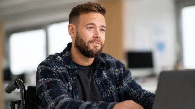 A young man in a wheelchair focused on his laptop in a contemporary office setting. The atmosphere is professional and inclusive.
