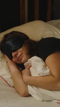 Vertical video. Woman wearing a sleep mask is sleeping in her bed. Person who sleeps peacefully and deeply.