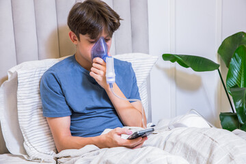 Teenage boy sitting in bed with a nebulizer inhaler on his face, holding a smartphone during inhalation therapy. Concept of home healthcare, asthma treatment, respiratory therapy
