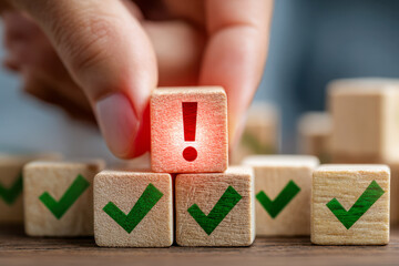 Hand placing a wooden block with a red exclamation mark above green checkmark blocks symbolizing important alerts or warnings amid approved tasks or decisions