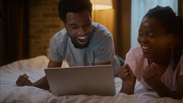 Happy excited African American woman girlfriend boyfriend man lying on bed family couple watching digital tablet shocked sport match game victory celebrating win shouting triumph at home bedroom night