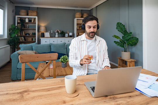 Young freelancer leading an online video meeting from a cozy home office smiling man with headphones gestures while speaking on a laptop during remote consulting and sharing project results.