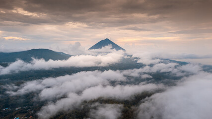 Epic aerial drone shot of Mount Inerie, a volcano on Flores, Indonesia. The peak rises above a sea of clouds, with a village and forest visible below.