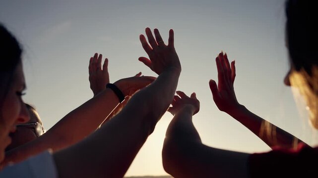 Hands reaching toward glowing sunset sky friend silhouette with palm raised in unity and joy while sun flare frames group gesture during outdoor play reaching contact and shared smiling expression