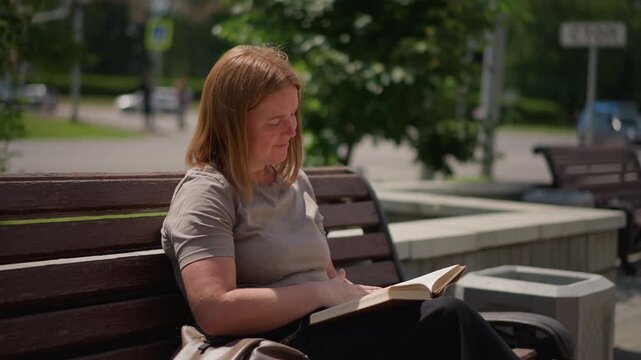 Student sitting on wooden bench studying book and flipping page under bright sunlight, calm expression and focused mind surrounded by green trees and soft background