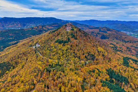Aerial view of a majestic mountain peak adorned with autumn's golden hues, crowned by ancient ruins against a backdrop of distant peaks, Orschwiller, Grand Est, France.