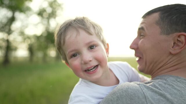 Man holding boy on shoulder in sunlit field smiling as father and son share warm embrace with child playful interaction and family bond visible in outdoor setting with green grass near oak tree