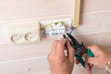 Electrician strips wire insulation while installing double wall socket on light wooden panel,...