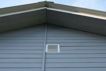 Blue wooden gable with white ventilation grille under pale sky, horizontal siding and roofline that...