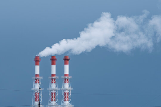 Three industrial chimneys release thick white smoke into the blue sky, symbolizing air pollution and environmental impact.