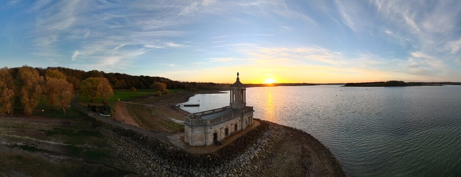 Aerial view of Normanton Church stands majestically against the vibrant sunset hues reflecting on the calm waters of Rutland Water, Oakham, England, United Kingdom.