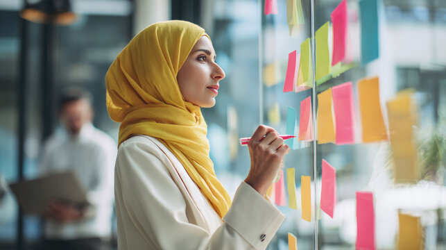 Confident businesswoman in yellow hijab writing on colorful sticky notes during brainstorming session in modern office, planning and organizing ideas for team project.