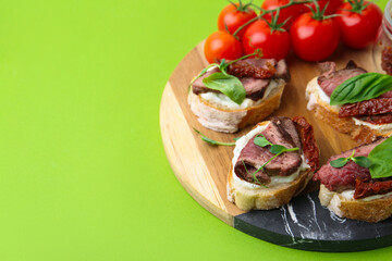 Bruschettas with roast beef, cream cheese, basil, sun-dried tomatoes and fresh vegetables on green background, closeup. Space for text
