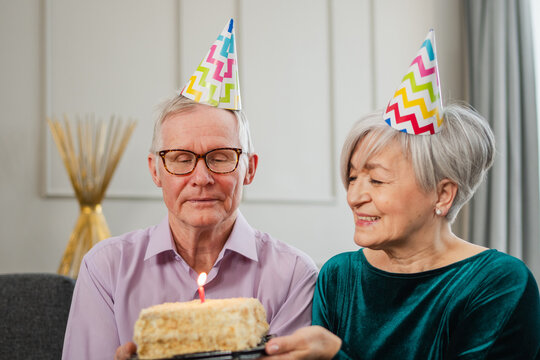 Make a wish. Family senior couple in party hat cap celebrating birthday anniversary together at home. Old man blowing out burning candles on birthday cake. Old woman wishes husband happy birthday - Powered by Adobe