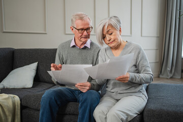 Sad tired disappointed middle aged senior couple sit with paper document. Unhappy older mature man woman reading paper bill managing bank finances calculating taxes planning loan debt pension payment