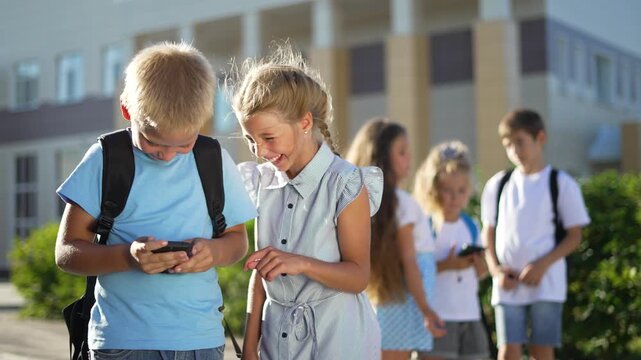 Children share smartphone at school entrance child boy and girl with backpack smile while student friend checks smartphone outdoor group of kid laughing near schoolyard building under sunlight